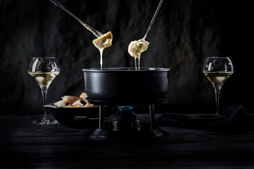 Zwei Stücke Brot eingetaucht in heisses Fäden ziehendes Käsefondue vom Chäsbueb und im Hintergrund Gläser Weisswein. Passend zum Fondue.